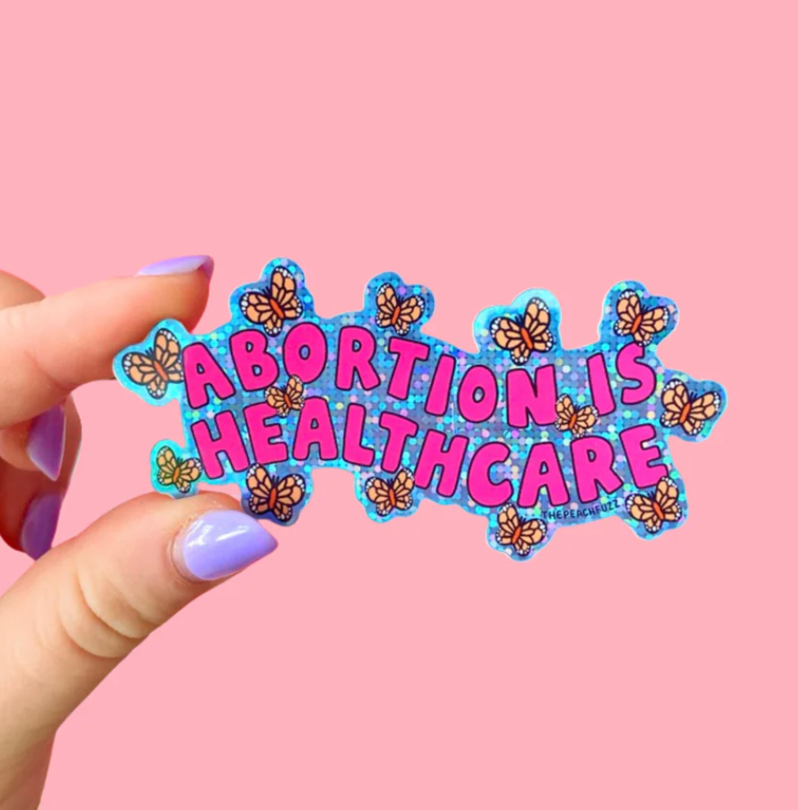Sticker with pink bubble letters saying "Abortion is healthcare" on a blue holographic glitter background surrounded by orange butterflies
