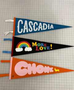 Three pennant flags in a row a blue one saying "Cascadia", a black one saying "More Love!", and an orange one saying "Chonk" 