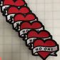 Red heart patch with a white banner across the heart that says "No one" in black letters